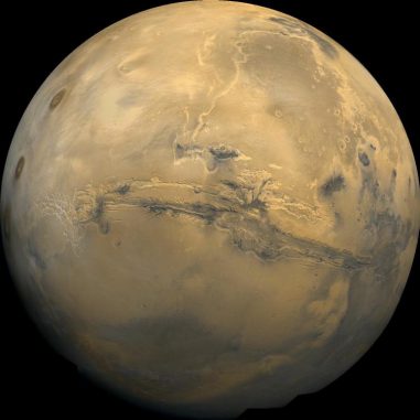 Der Mars. In der Bildmitte erkennt man das größte Canyonsystem des Sonnensystems, Valles Marineris. Die dunklen Flecken links sind die Vulkane Ascraeus Mons, Pavonis Mons und Arsia Mons. Vulkanische Aktivität könnte in der Frühzeit des Planeten genug Methan freigesetzt haben, um wärmere Bedingungen zu schaffen, die flüssiges Wasser auf der Oberfläche ermöglichten. (Jody Swann / Tammy Becker / Alfred McEwen / USGS / NASA)