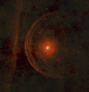 Beteigeuze (Mitte) ist von staubigen Materiebögen umgeben, die der Stern im Laufe seiner Entwicklung zu einem roten Superriesen abgestoßen hat. Die geradlinige Struktur links steht vermutlich mit dem galaktischen Magnetfeld in Zusammenhang, oder stellt den Rand einer interstellaren Wolke dar. (ESA / Herschel / PACS / L. Decin et al)